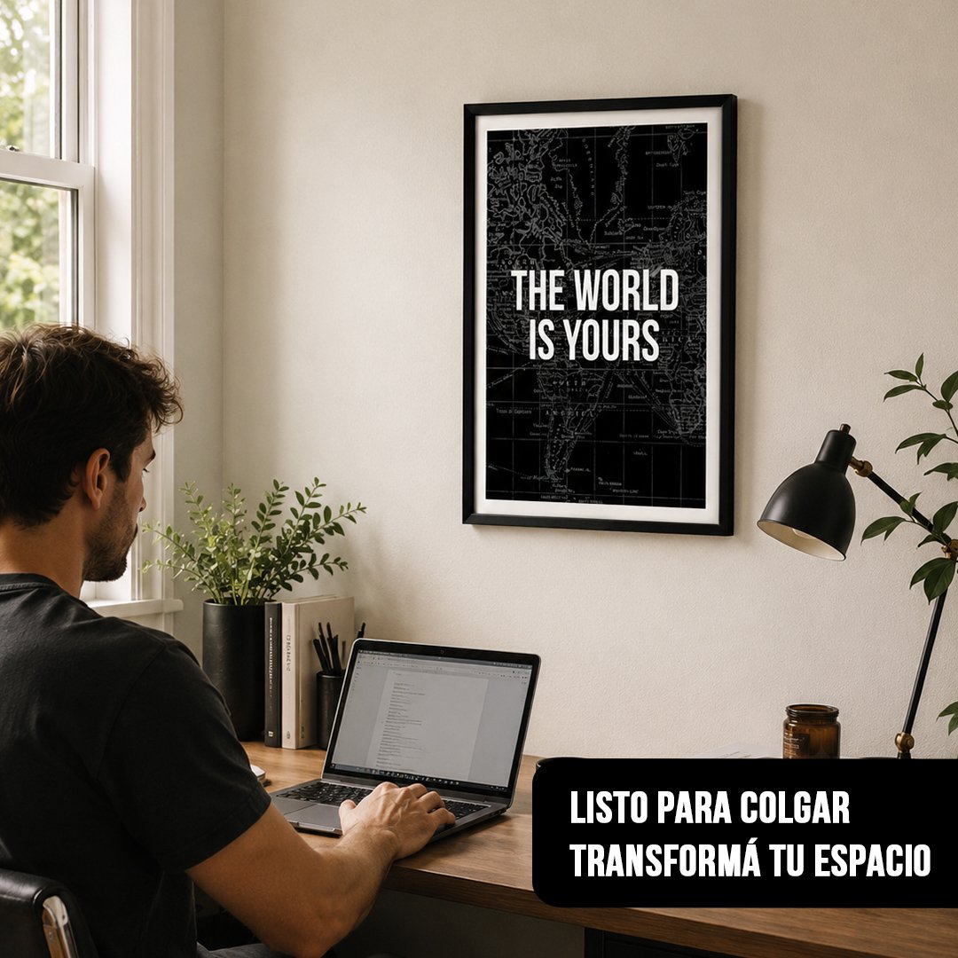 Cuadro vertical motivacional con frase "THE WORLD IS YOURS" sobre mapa negro enmarcado en negro, colgado sobre un escritorio de oficina con un hombre usando una laptop