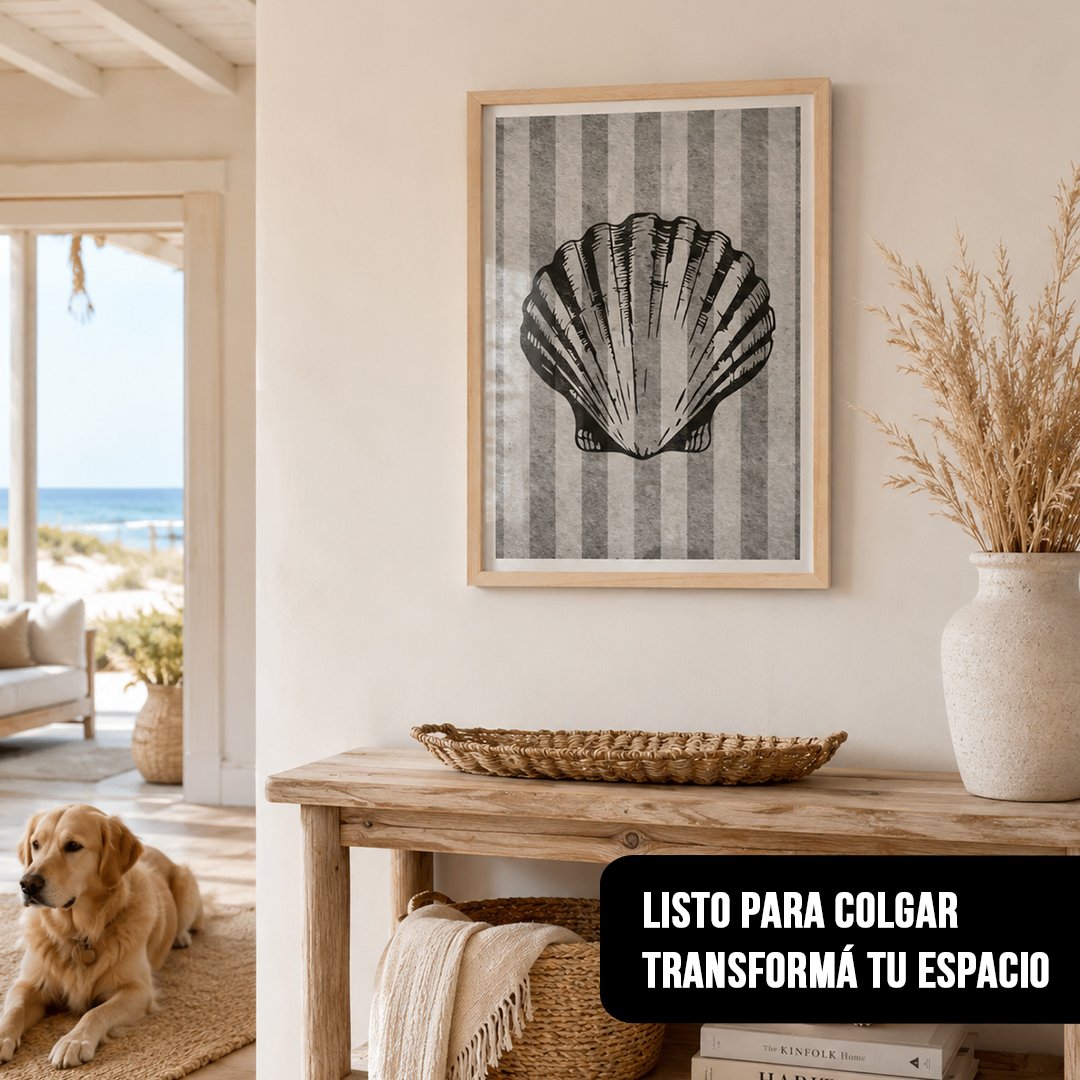 Cuadro vertical con marco de madera clara que muestra una concha marina negra sobre fondo rayado gris, colgado sobre una mesa rústica en un ambiente costero con un perro en el suelo