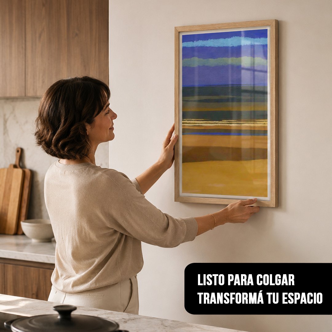 Cuadro vertical con marco de madera clara de un paisaje abstracto en tonos violetas y ocres, sostenido por una mujer en una cocina moderna