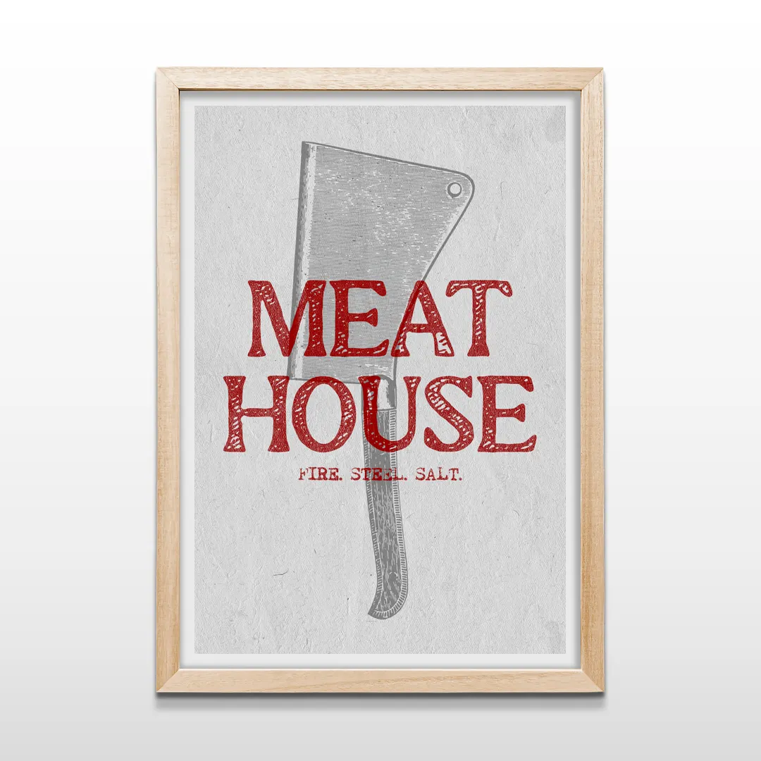 Cuadro vertical enmarcado en madera clara con ilustración de hacha de carnicero y el texto "MEAT HOUSE FIRE STEEL SALT"