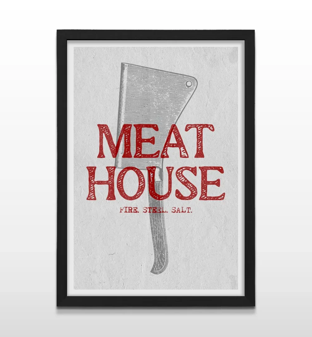 Cuadro vertical enmarcado en negro mate con ilustración de hacha de carnicero y leyenda "MEAT HOUSE FIRE STEEL SALT"