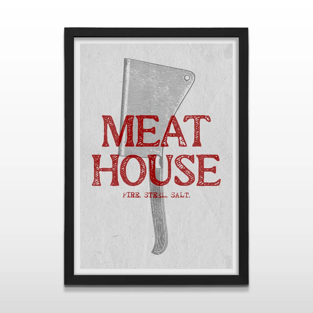 Cuadro vertical enmarcado en negro mate con ilustración de hacha de carnicero y leyenda "MEAT HOUSE FIRE STEEL SALT"
