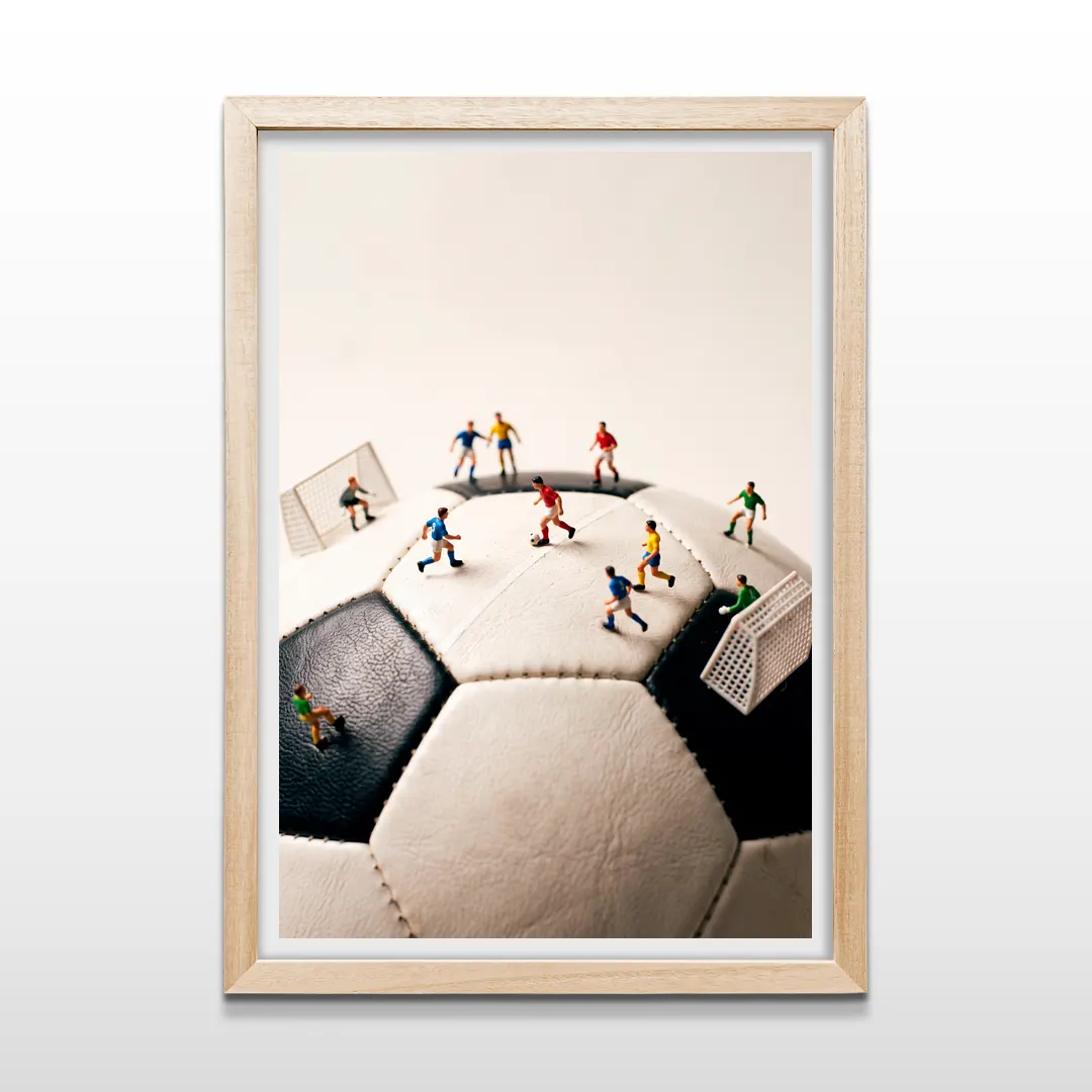 Cuadro vertical en marco de madera clara con foto macro de partido de fútbol diminuto montado sobre pelota tradicional